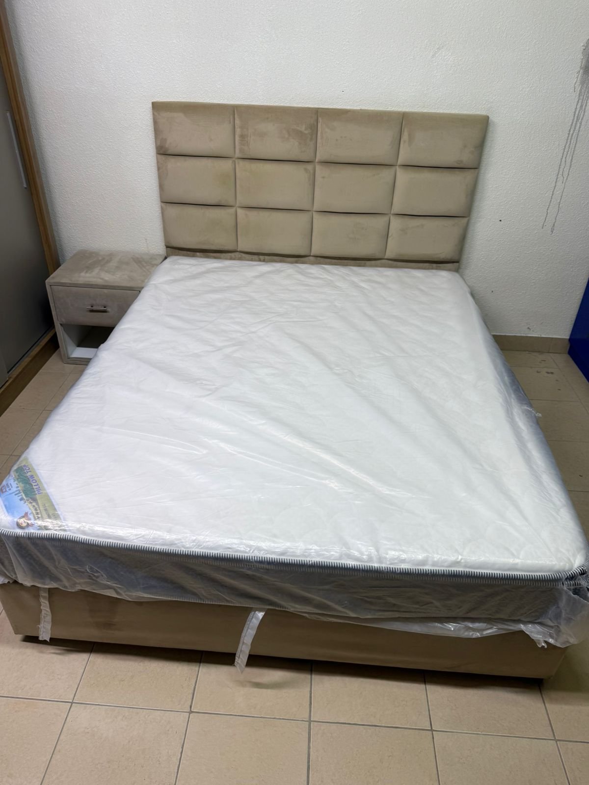 The beige upholstered bed features a checkered, padded headboard and comes with a new mattress.