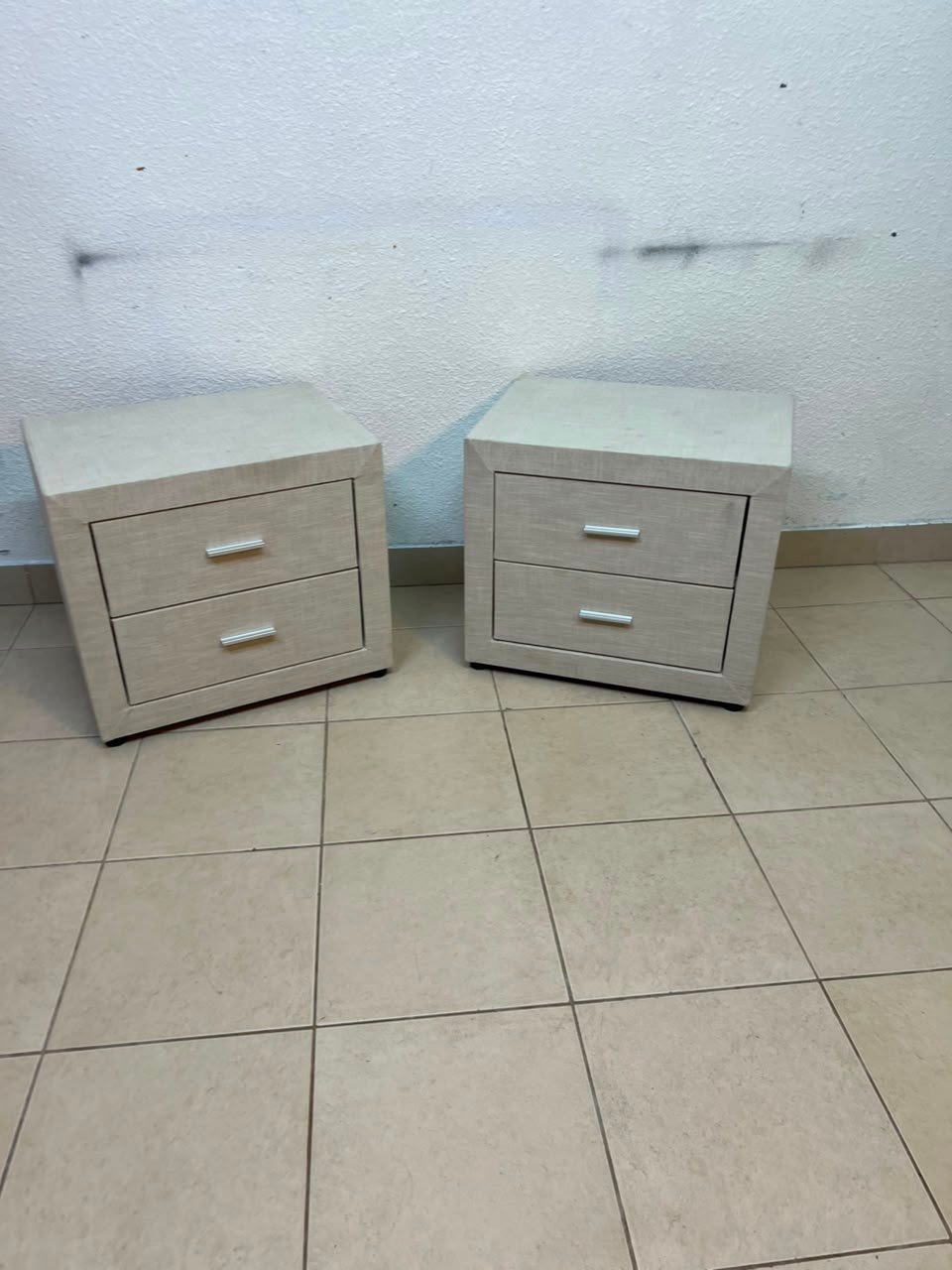 A beige upholstered nightstand with two drawers and silver handles.