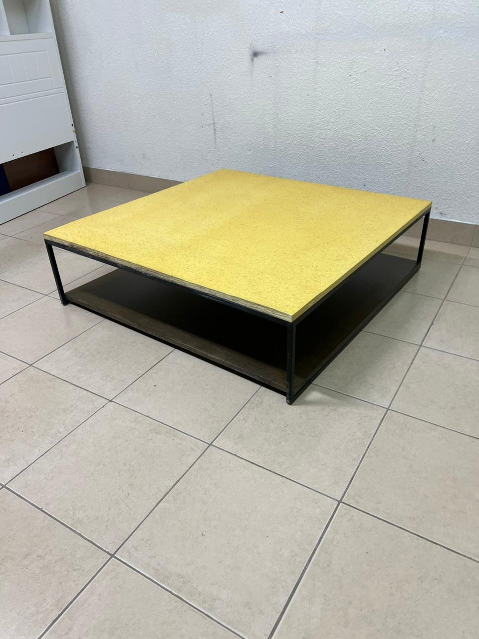 This is a square coffee table with an industrial design, featuring a black metal frame, a black pull-out drawer or lower shelf, and a light yellow wooden top.