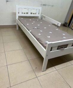 A white wooden cot, believed to be the KRITTER model from IKEA UAE, measuring approximately 70 x 160 cm, and comes with a side guard rail