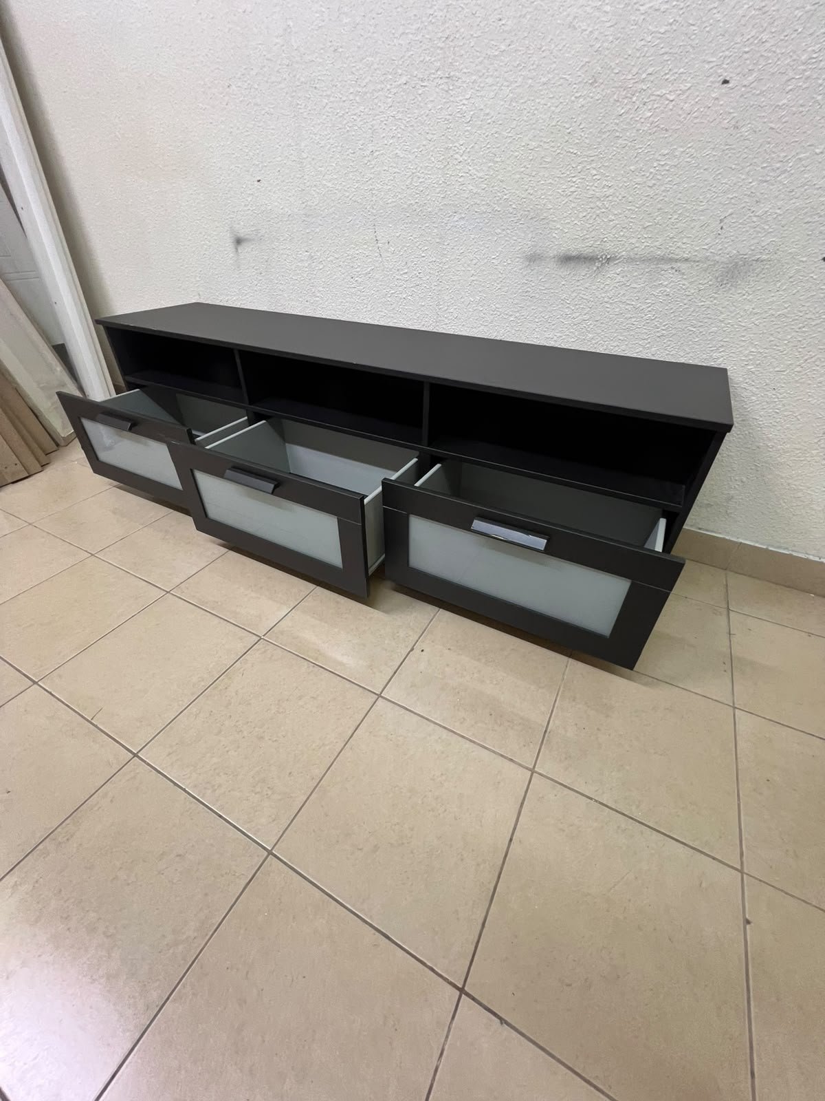 The television unit shown in the picture is a modern black TV stand with three drawers featuring opaque glass fronts and open shelves for appliances. - Image 4
