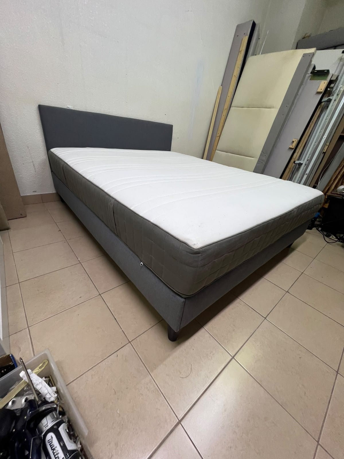 A gray upholstered bed frame with a white mattress and blanket, or a dark gray mattress on the side.