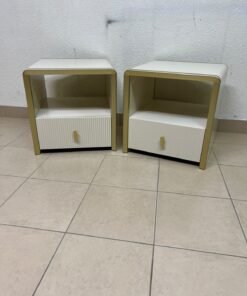 The image shows two modern bedside tables in a creamy white finish, featuring gold frames and details, as well as ribbed drawer fronts (from Home Centre).