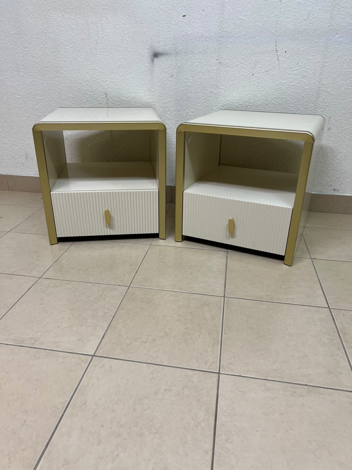 The image shows two modern bedside tables in a creamy white finish, featuring gold frames and details, as well as ribbed drawer fronts (from Home Centre).