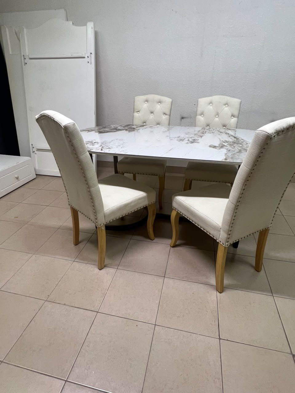 An oval dining table with a marble-patterned ceramic top, surrounded by four chairs upholstered in light beige fabric and featuring button-tufted backrests.