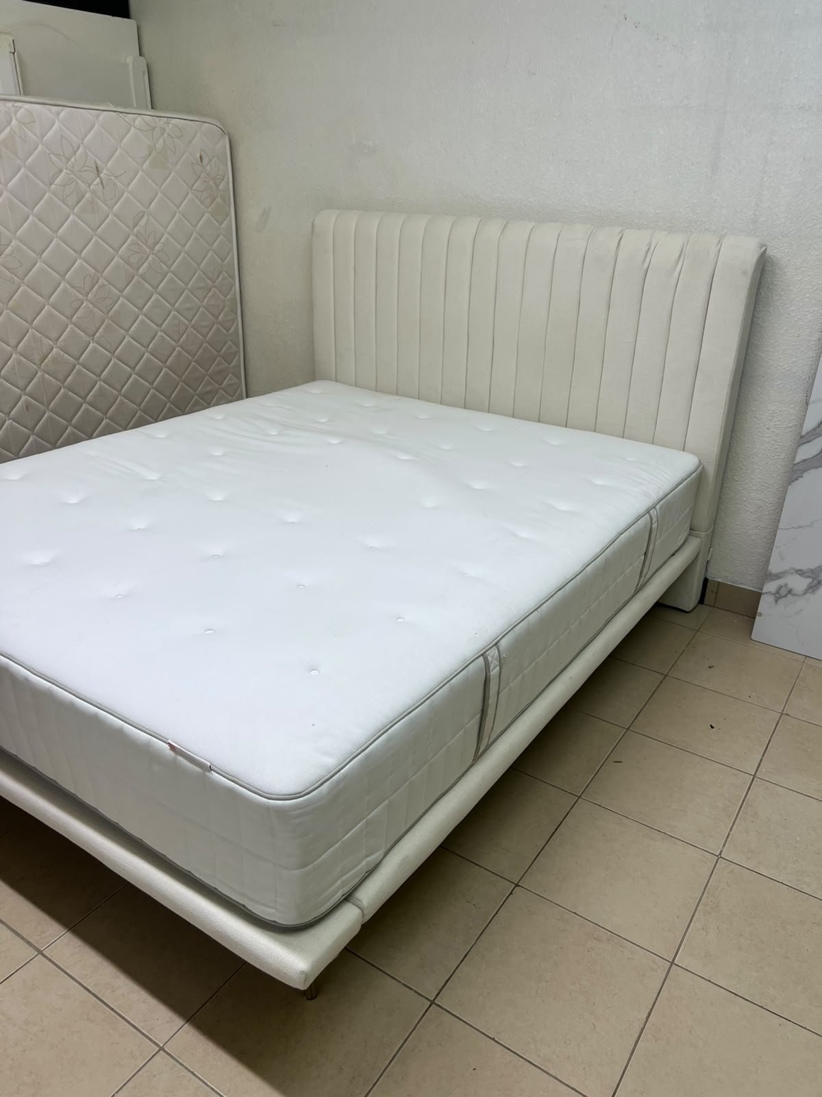 An upholstered double bed in white or light beige with a striped headboard, featuring a comfortable white mattress – Home Centre.