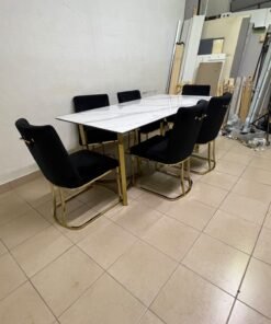 A modern six-seater dining set featuring an elegant design that combines a white marble-patterned tabletop with a gold metal base and frame, paired with black velvet chairs featuring matching gold legs.
