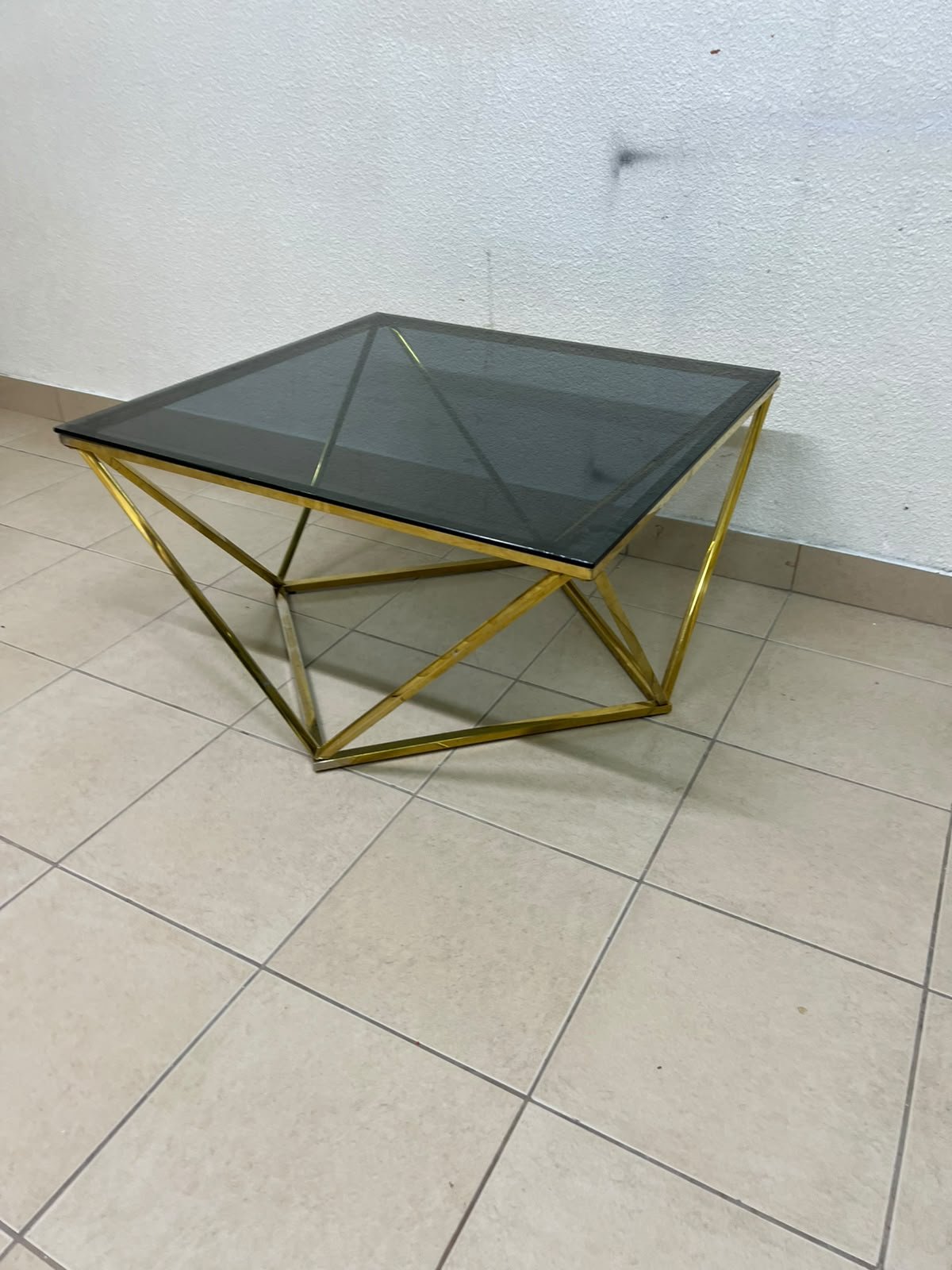 A modern, square coffee table featuring a dark or smoked glass top and a cohesive, diamond-inspired geometric base made of metal or polished gold-tone steel.