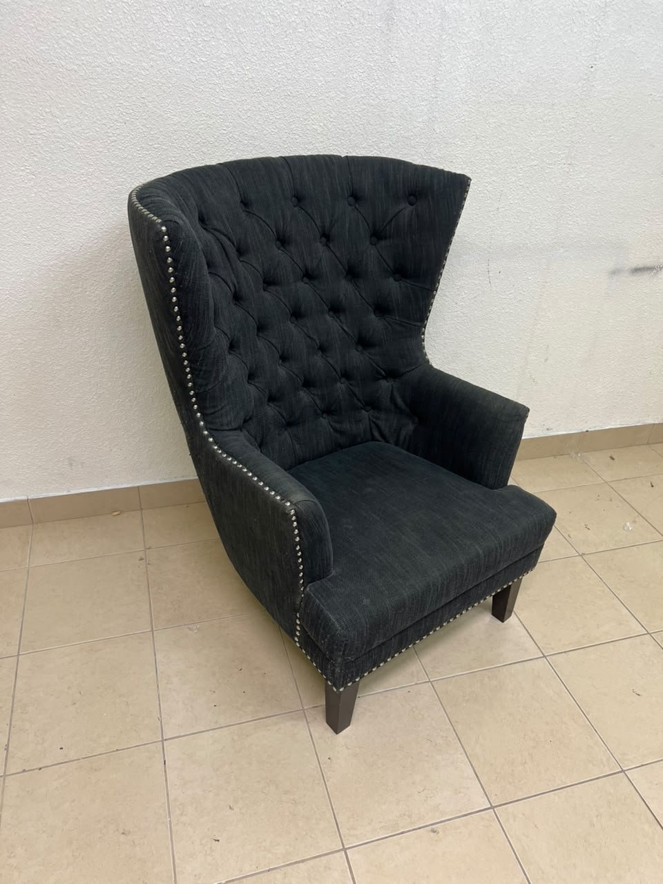 A single upholstered wingback chair featuring a button-tufted back, a black or dark gray fabric finish, and metal nailhead trim along the edges.