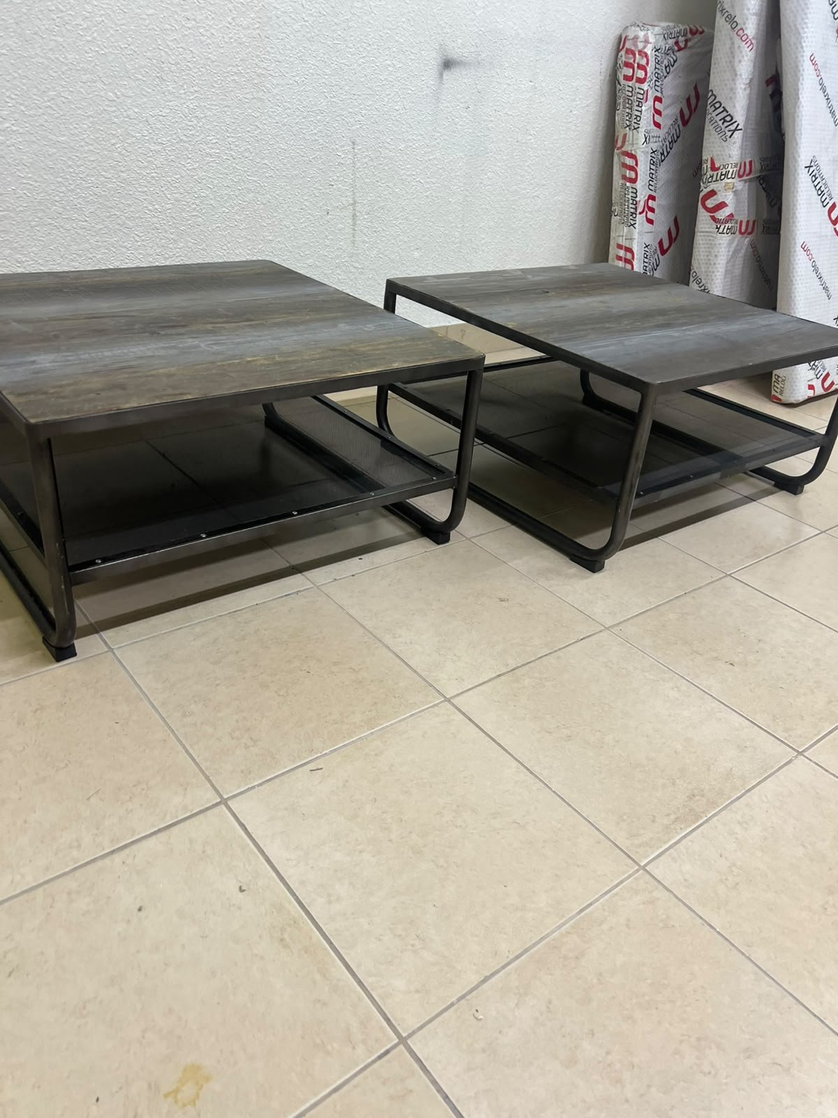 A set of two coffee tables featuring a modern industrial design; each boasts a wooden tabletop with a rustic or distressed look, a sturdy black metal frame and base, and a practical bottom shelf.