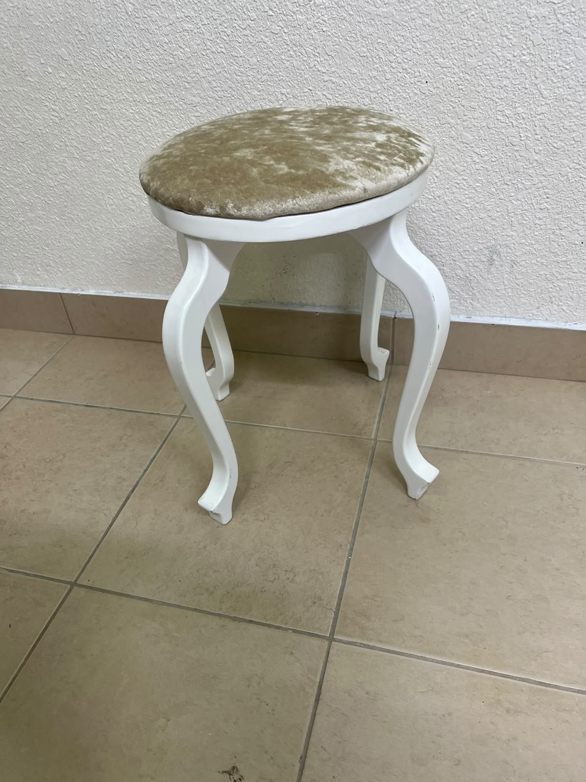 A vanity stool featuring a classic French or "Queen Anne" design, characterized by a white wooden frame with cabriole legs and a round seat upholstered in beige or light-gold velvet fabric.