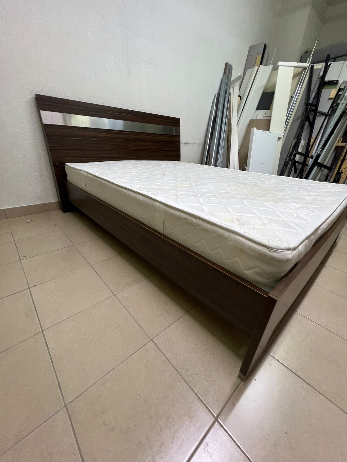 A dark brown wooden double bed featuring a headboard accented with a silver metal strip, and equipped with a white padded mattress.