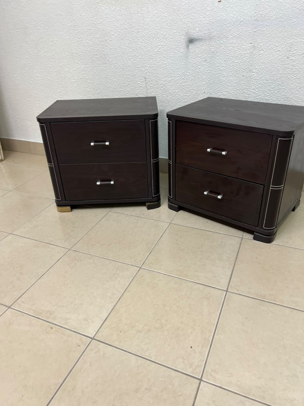 Two dark brown wooden bedside tables (nightstands) featuring two drawers and curved corners, accented with contrasting white stitching and silver-colored metal drawer handles.