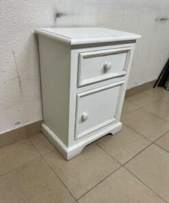 A white side table or nightstand featuring a traditional, classic design, comprising an upper drawer and a lower cabinet with a door, both fitted with round wooden knobs.
