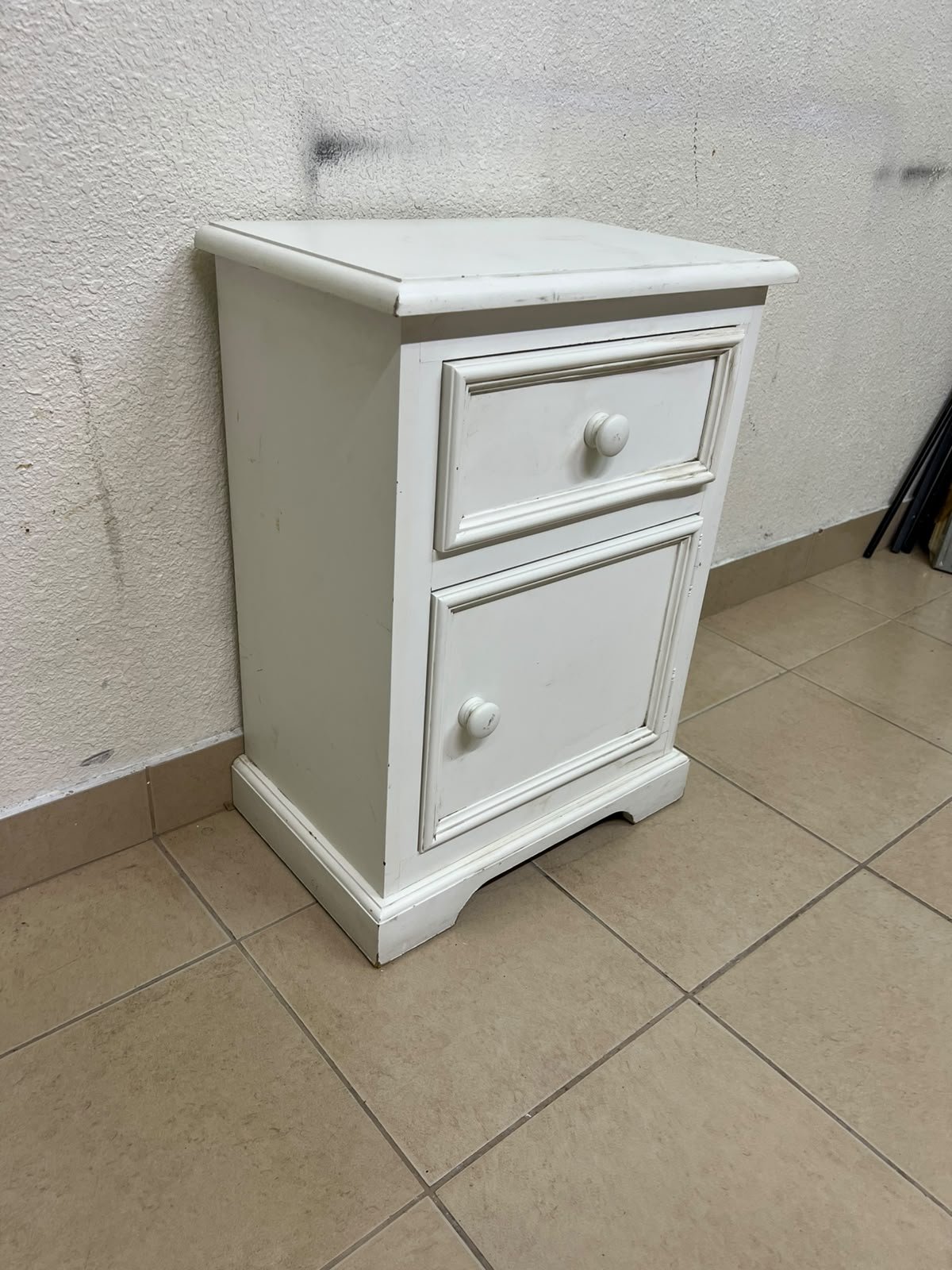 A white side table or nightstand featuring a traditional, classic design, comprising an upper drawer and a lower cabinet with a door, both fitted with round wooden knobs.
