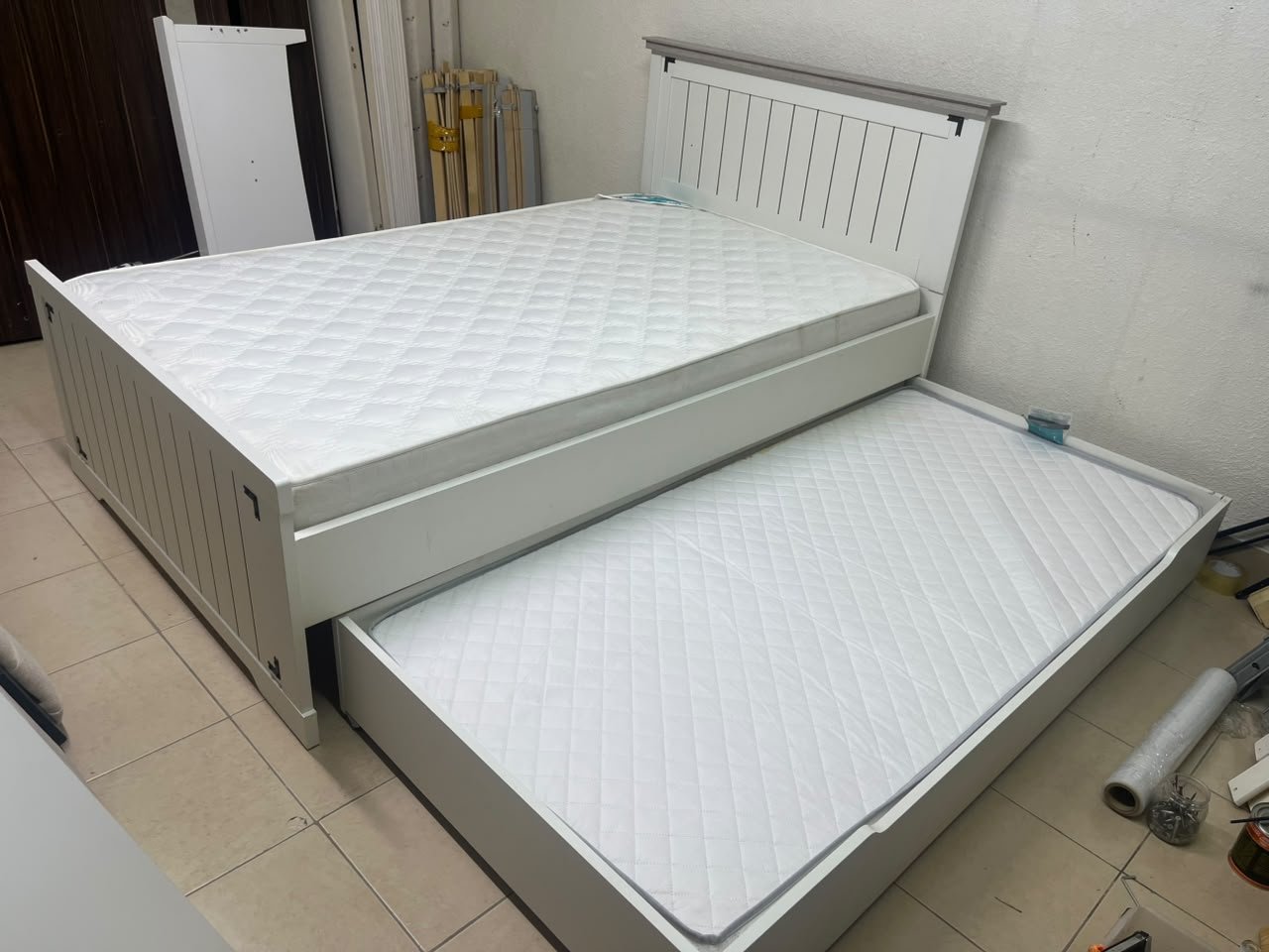 A white wooden single bed featuring a pull-out trundle bed underneath, with two matching padded mattresses.