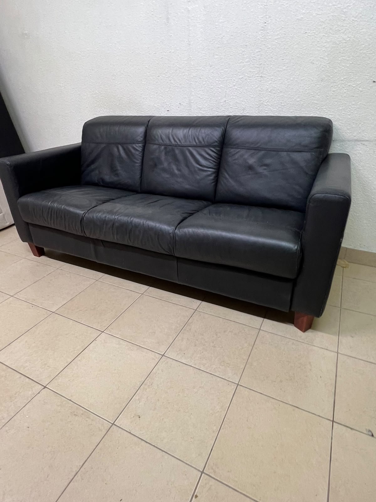 A modern three-seater black leather sofa featuring square armrests and wooden legs, resembling the IKEA "Skogaby" model.