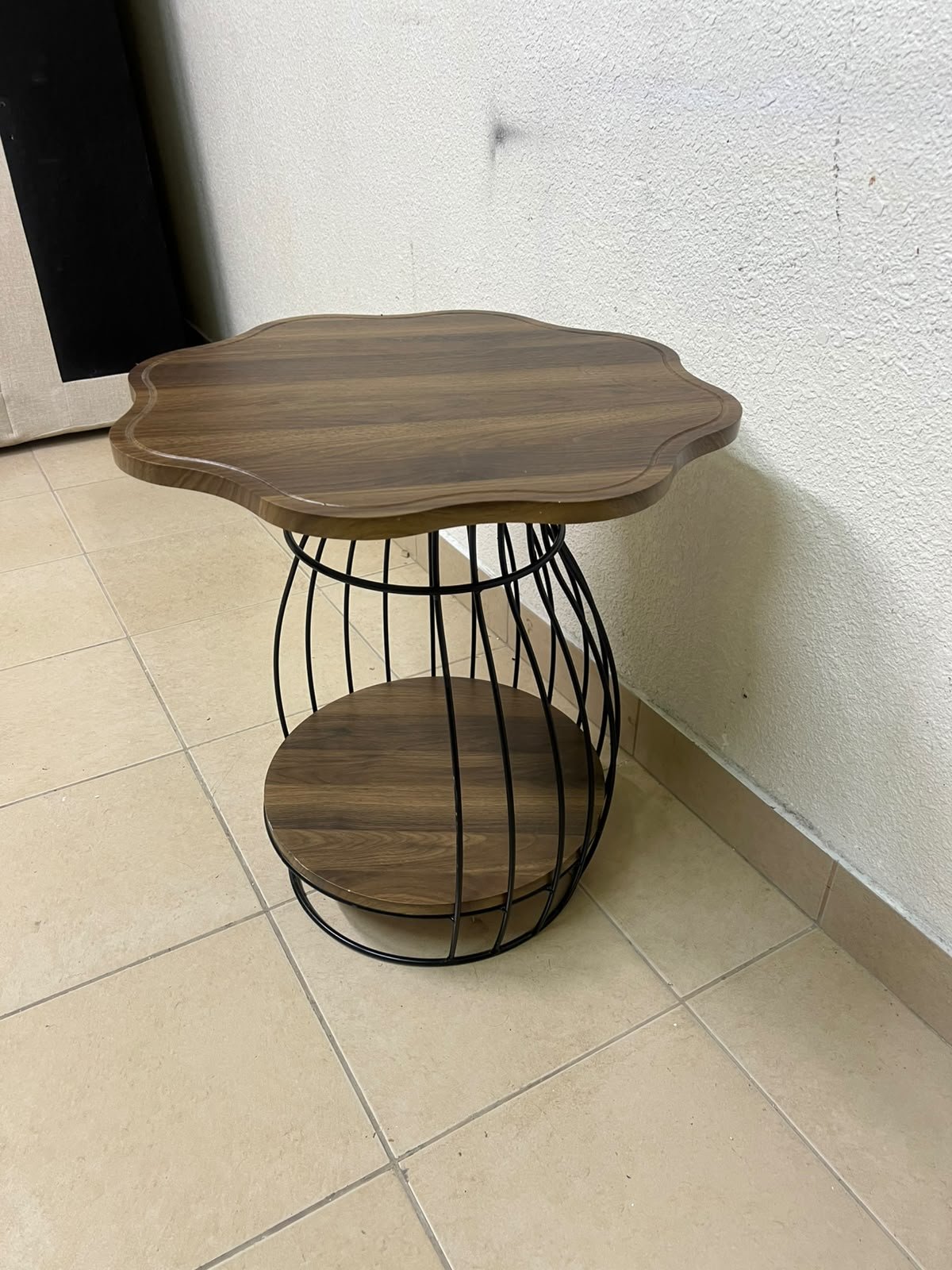 This modern two-tier side table features a wooden top with curved edges and a lower storage shelf enclosed by a black metal frame designed like a wire mesh cage.