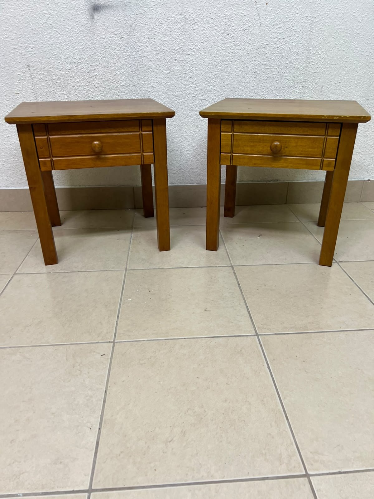 Two classic, medium-toned wooden side tables featuring a single drawer with a round wooden knob and classic grooved detailing on the frame.