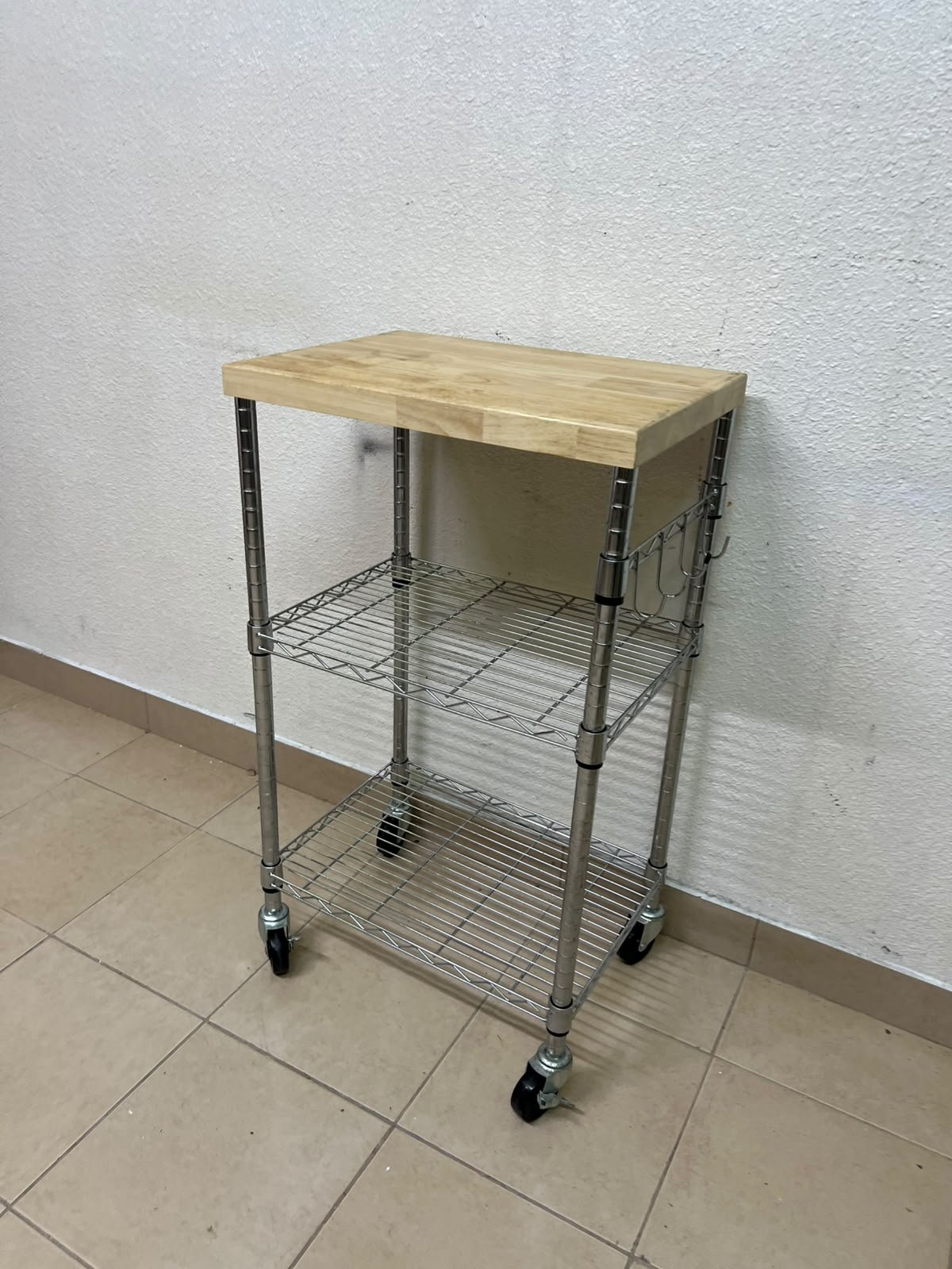 A multi-shelf kitchen cart made of chrome wire, featuring a light-colored wood top (butcher block style) and swivel casters.