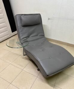 A modern grey faux leather chaise longue featuring an attached round glass side table and metal legs.
