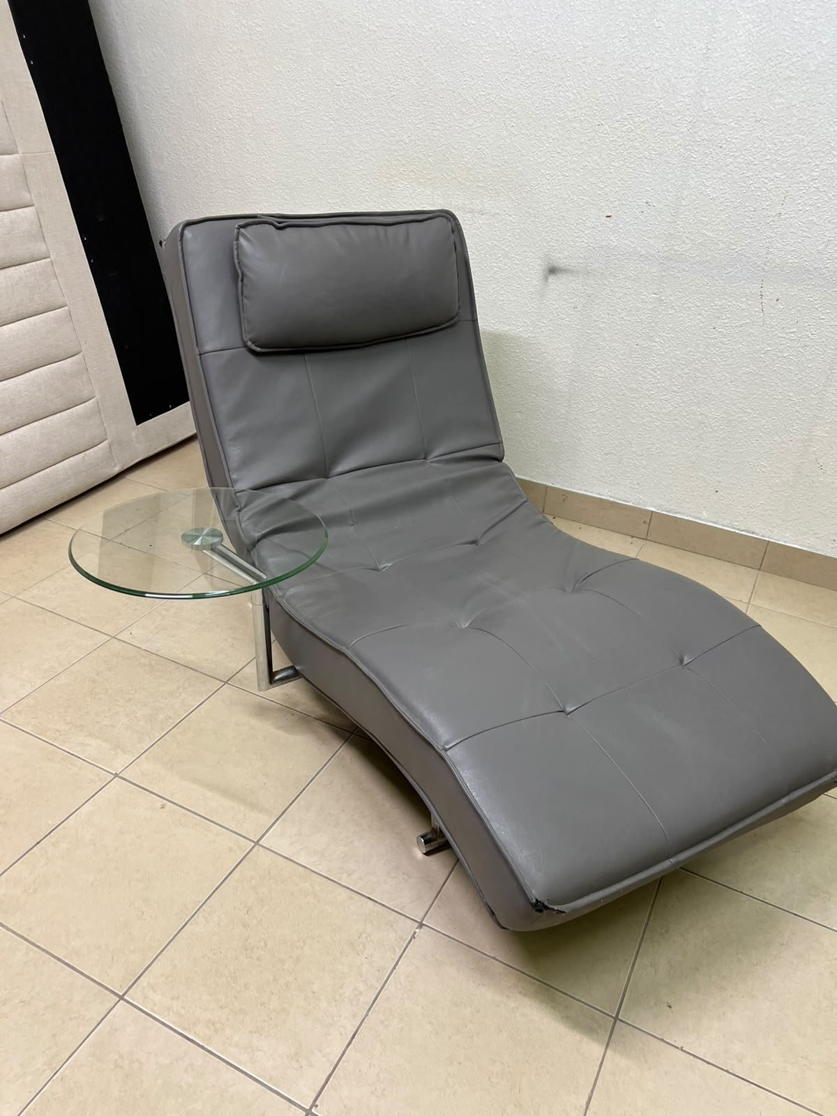 A modern grey faux leather chaise longue featuring an attached round glass side table and metal legs.