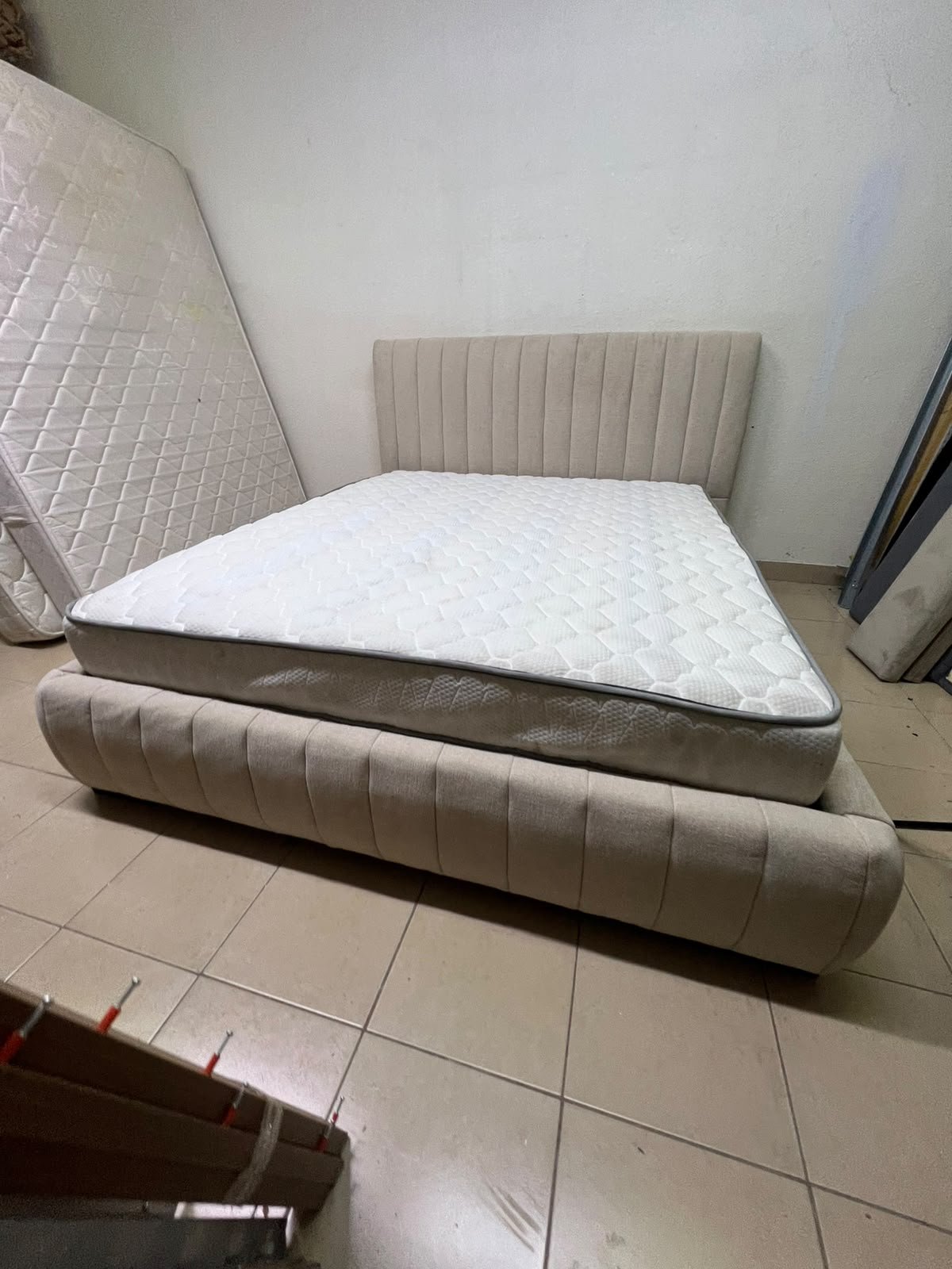 A bed featuring a modern design and a beige upholstered frame, complete with a headboard designed with padded vertical channels and a matching upholstered base; its style resembles the "Curvy Haze" upholstered bed available at Home Box. - Image 5