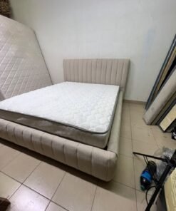 A bed featuring a modern design and a beige upholstered frame, complete with a headboard designed with padded vertical channels and a matching upholstered base; its style resembles the "Curvy Haze" upholstered bed available at Home Box.