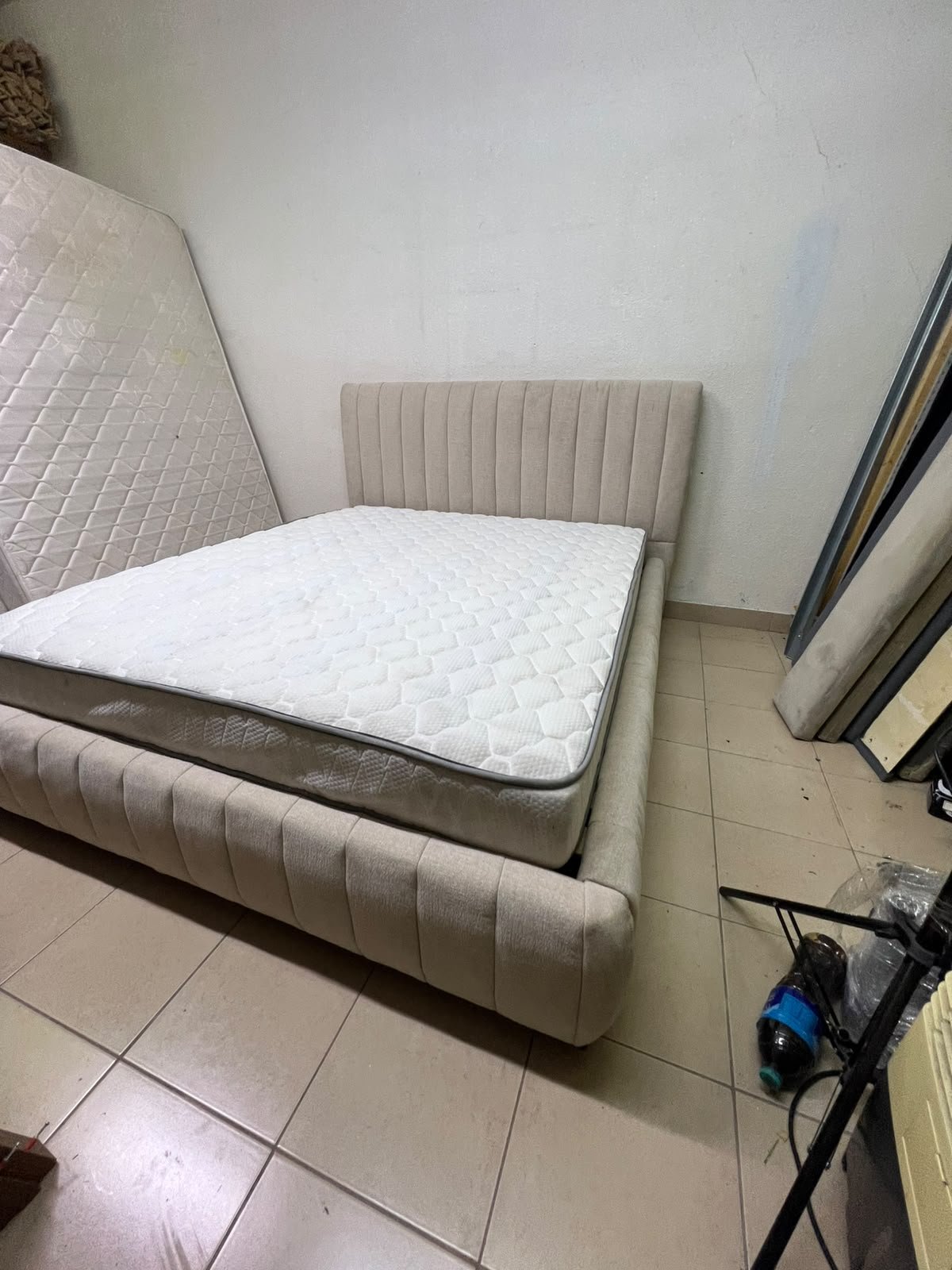 A bed featuring a modern design and a beige upholstered frame, complete with a headboard designed with padded vertical channels and a matching upholstered base; its style resembles the "Curvy Haze" upholstered bed available at Home Box.