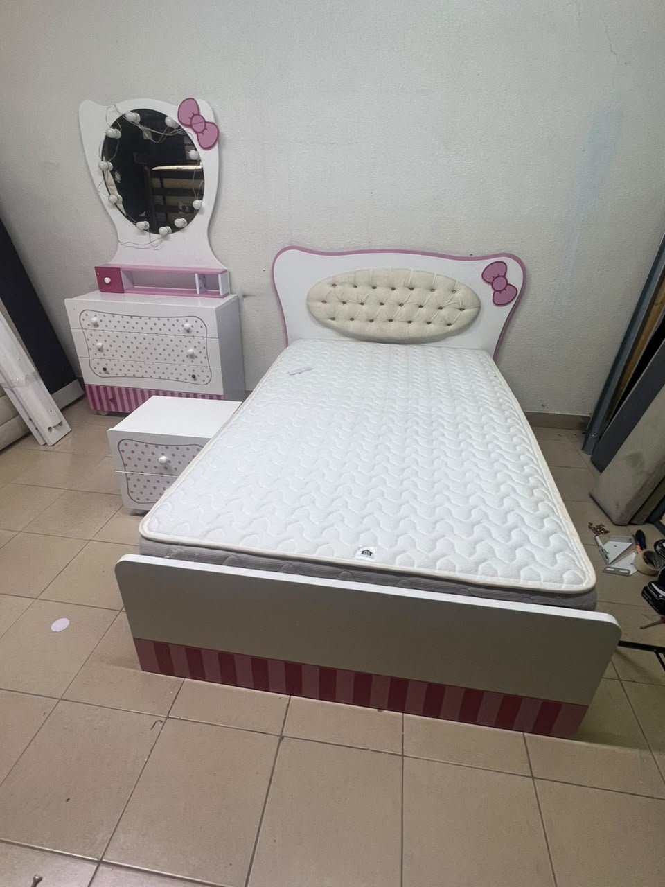 A girls' bedroom featuring a white and pink color scheme, including a bed with bedding, a storage table with an illuminated mirror, and a bedside table.