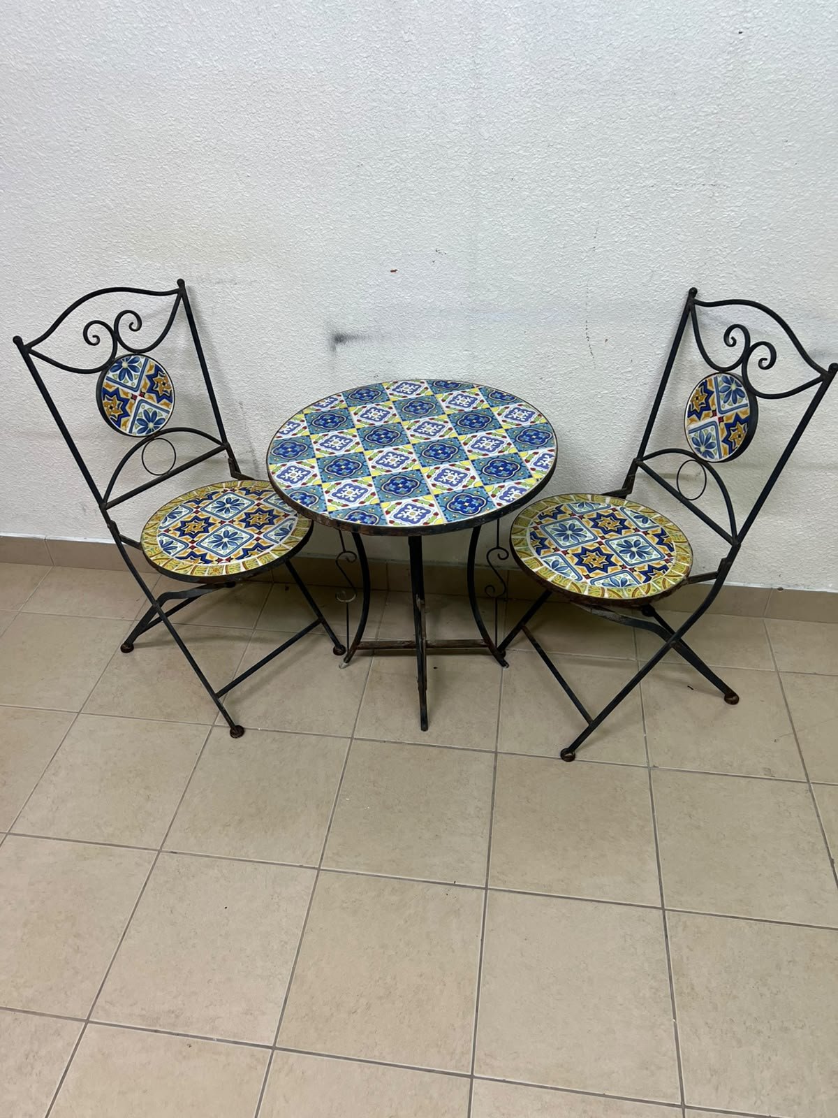 A three-piece black wrought-iron table and chair set, featuring blue and yellow ceramic mosaic tile accents on the round tabletop and the seats of the foldable chairs. - Image 2
