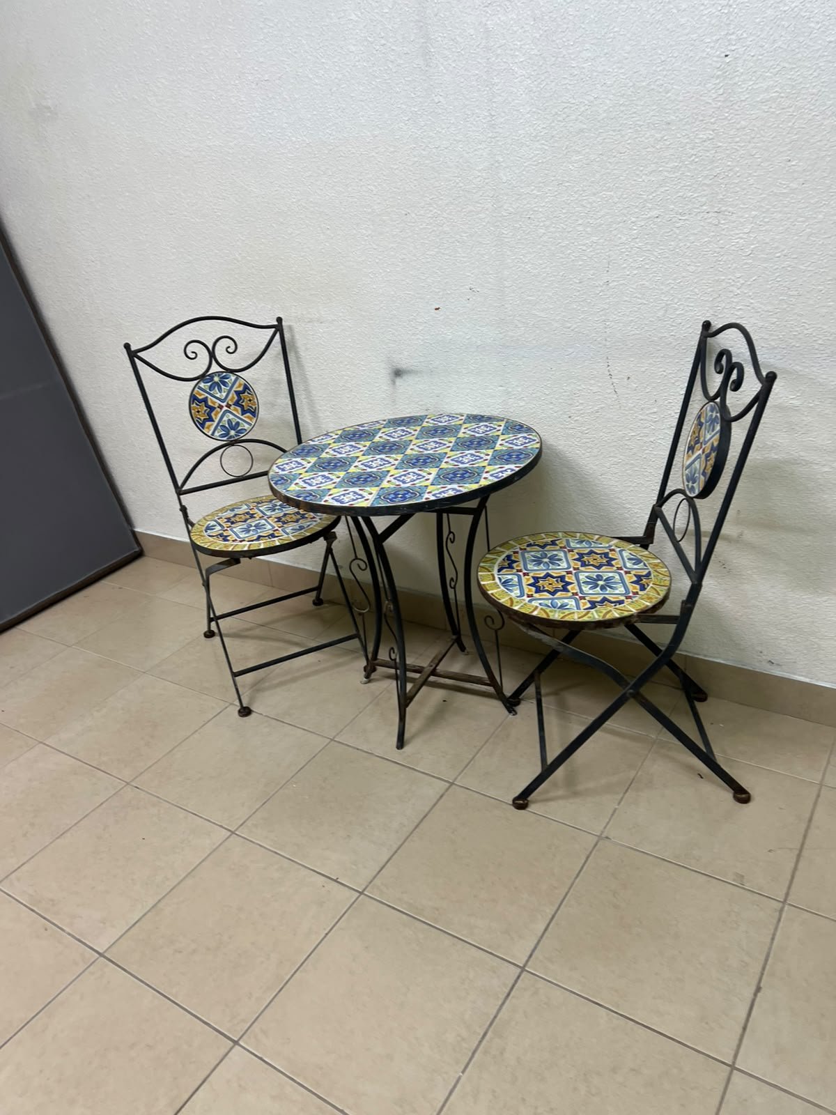 A three-piece black wrought-iron table and chair set, featuring blue and yellow ceramic mosaic tile accents on the round tabletop and the seats of the foldable chairs. - Image 3
