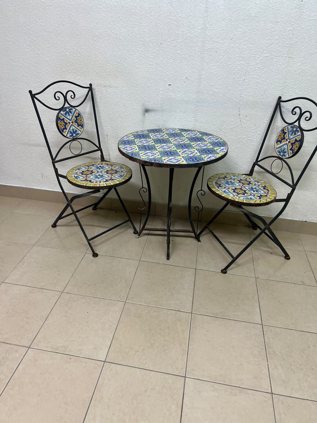 A three-piece black wrought-iron table and chair set, featuring blue and yellow ceramic mosaic tile accents on the round tabletop and the seats of the foldable chairs. - Image 4