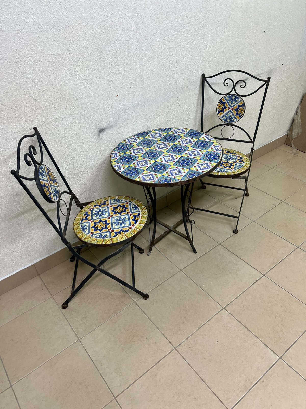 A three-piece black wrought-iron table and chair set, featuring blue and yellow ceramic mosaic tile accents on the round tabletop and the seats of the foldable chairs.