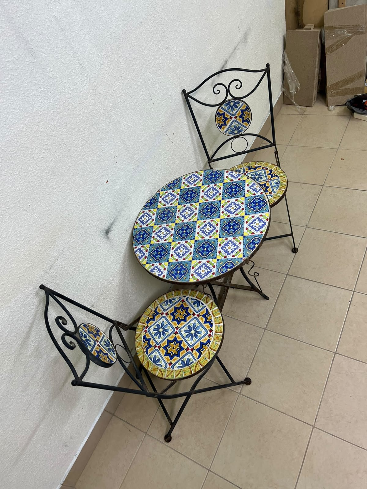 A three-piece black wrought-iron table and chair set, featuring blue and yellow ceramic mosaic tile accents on the round tabletop and the seats of the foldable chairs. - Image 9