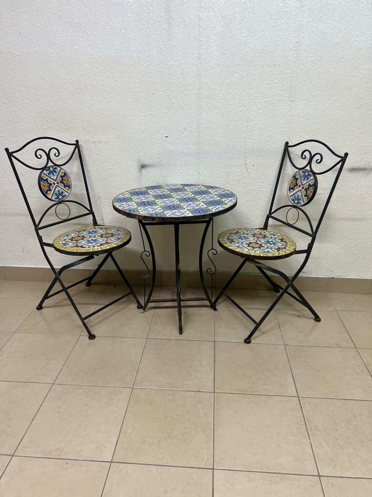 A three-piece black wrought-iron table and chair set, featuring blue and yellow ceramic mosaic tile accents on the round tabletop and the seats of the foldable chairs. - Image 11
