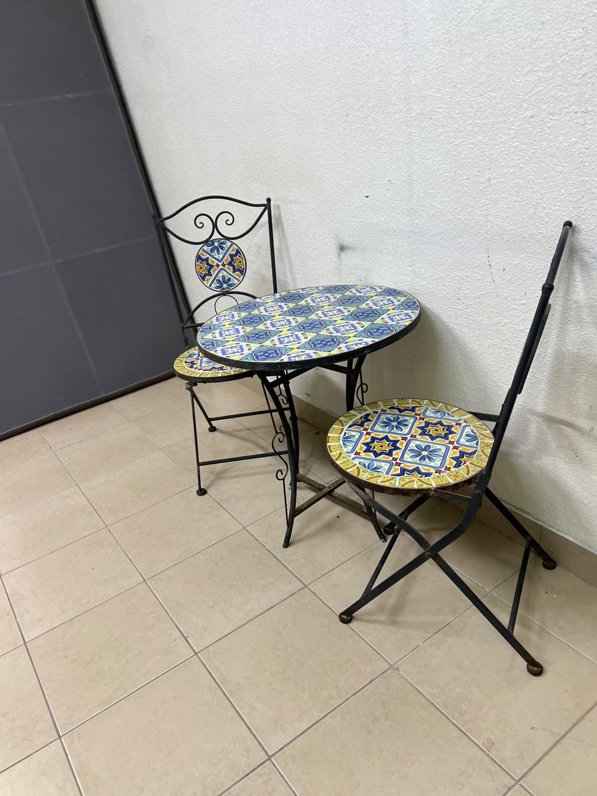 A three-piece black wrought-iron table and chair set, featuring blue and yellow ceramic mosaic tile accents on the round tabletop and the seats of the foldable chairs. - Image 5