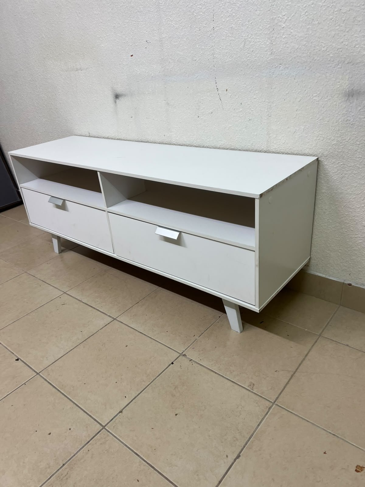 A modern, white, freestanding TV stand featuring angled legs and two large lower storage drawers, topped by open shelves.