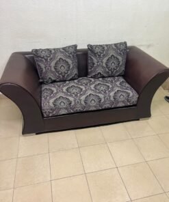 The sofa is designed for two people and upholstered in dark brown faux leather, with a purple and gray damask-patterned fabric covering the seat cushions and matching throw pillows.