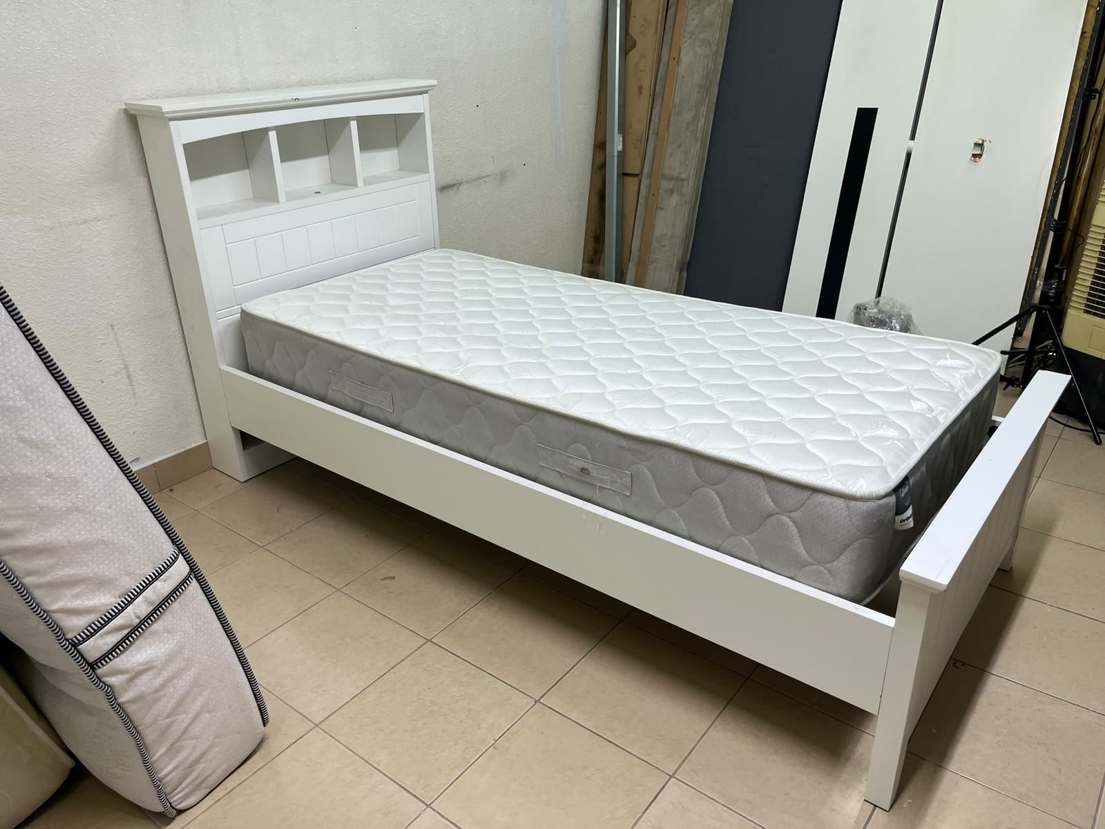 A white wooden single bed frame featuring a headboard with built-in bookshelves, and equipped with a standard padded mattress.