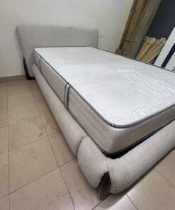 A modern, curved bed frame upholstered in off-white bouclé fabric with dark wood accents, complemented by a patterned, cushioned mattress.