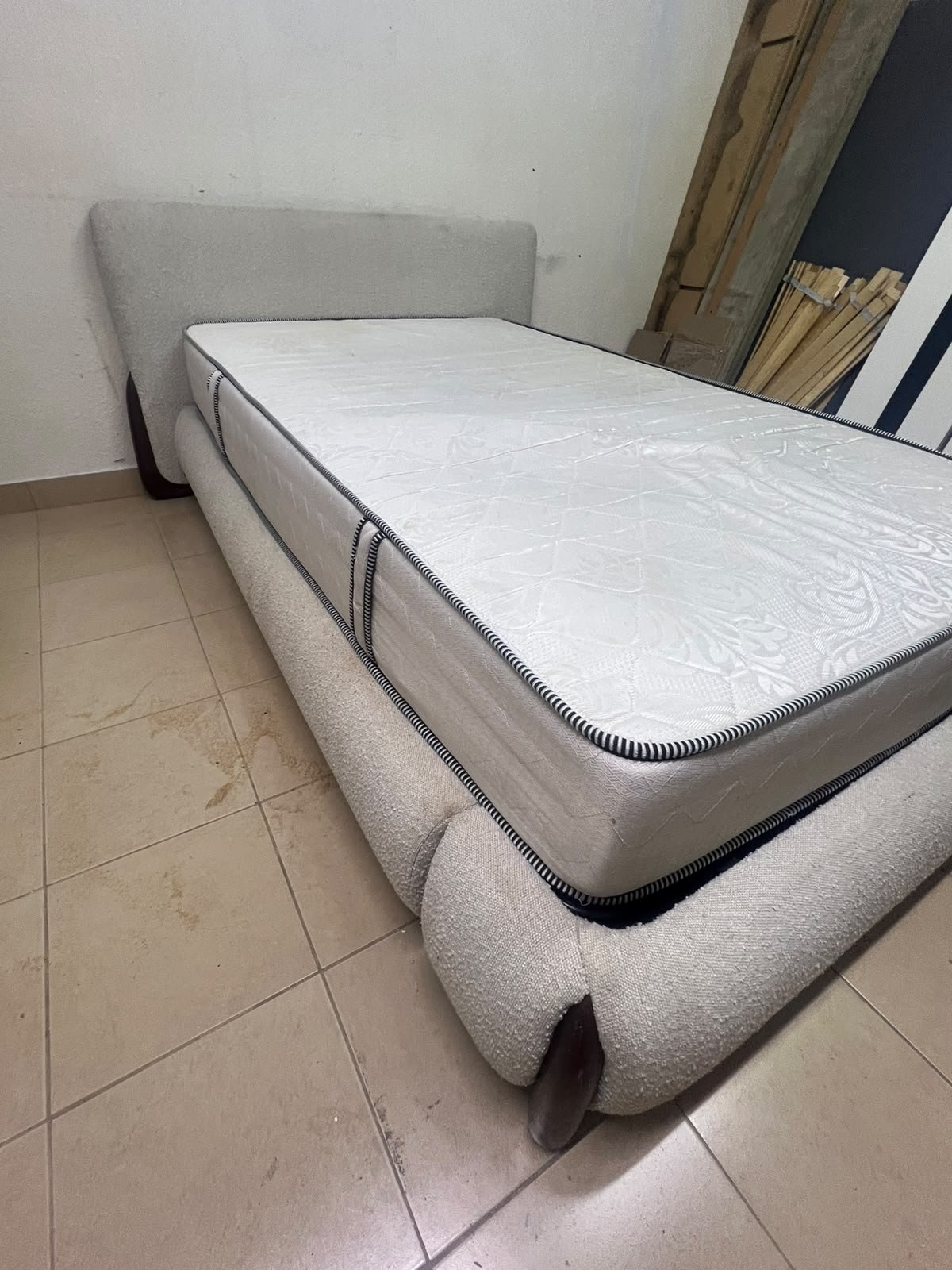 A modern, curved bed frame upholstered in off-white bouclé fabric with dark wood accents, complemented by a patterned, cushioned mattress.