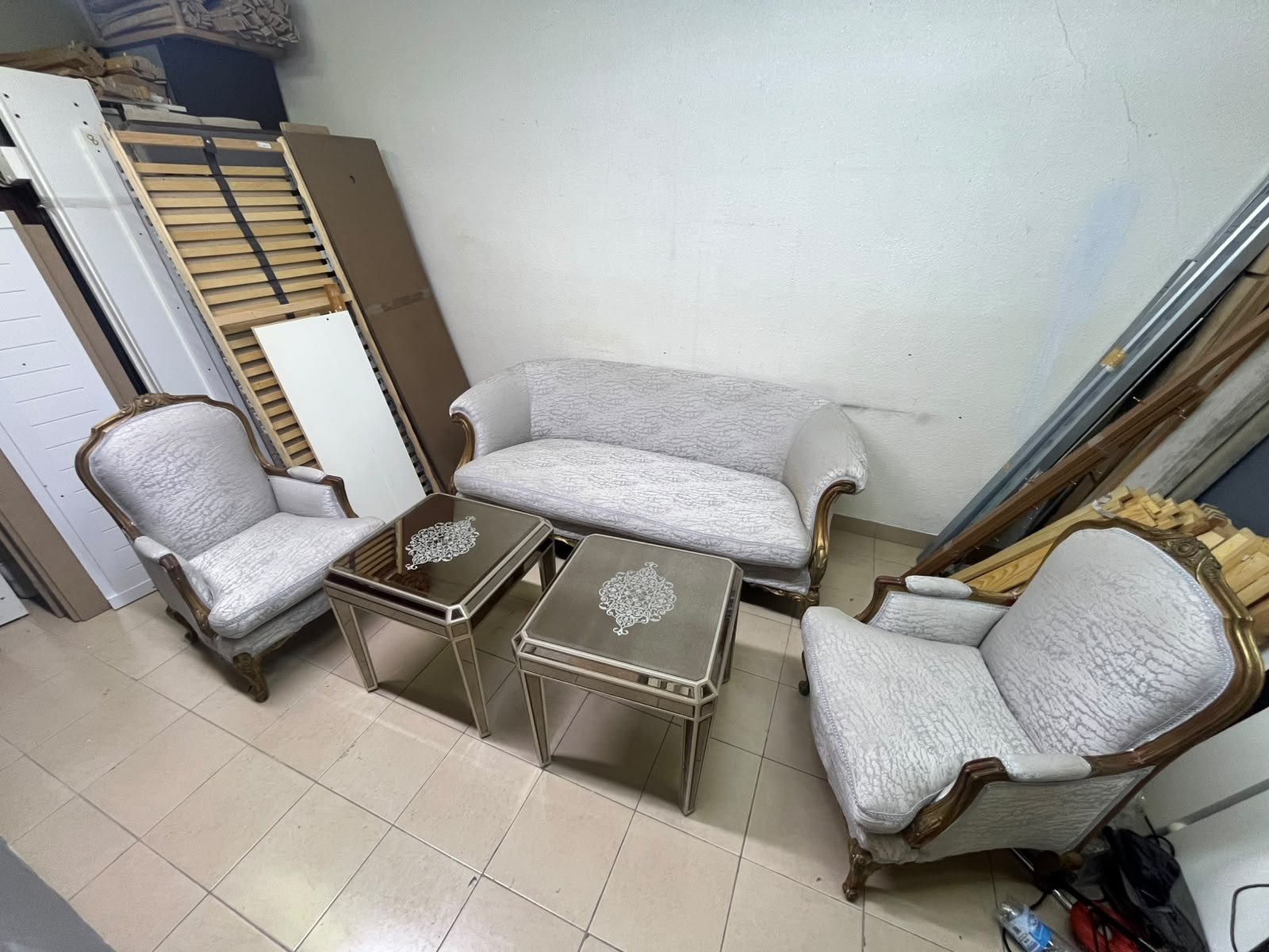 A light-colored, upholstered classic living room set featuring a sofa and armchairs with carved wooden frames, along with two matching mirrored side tables.