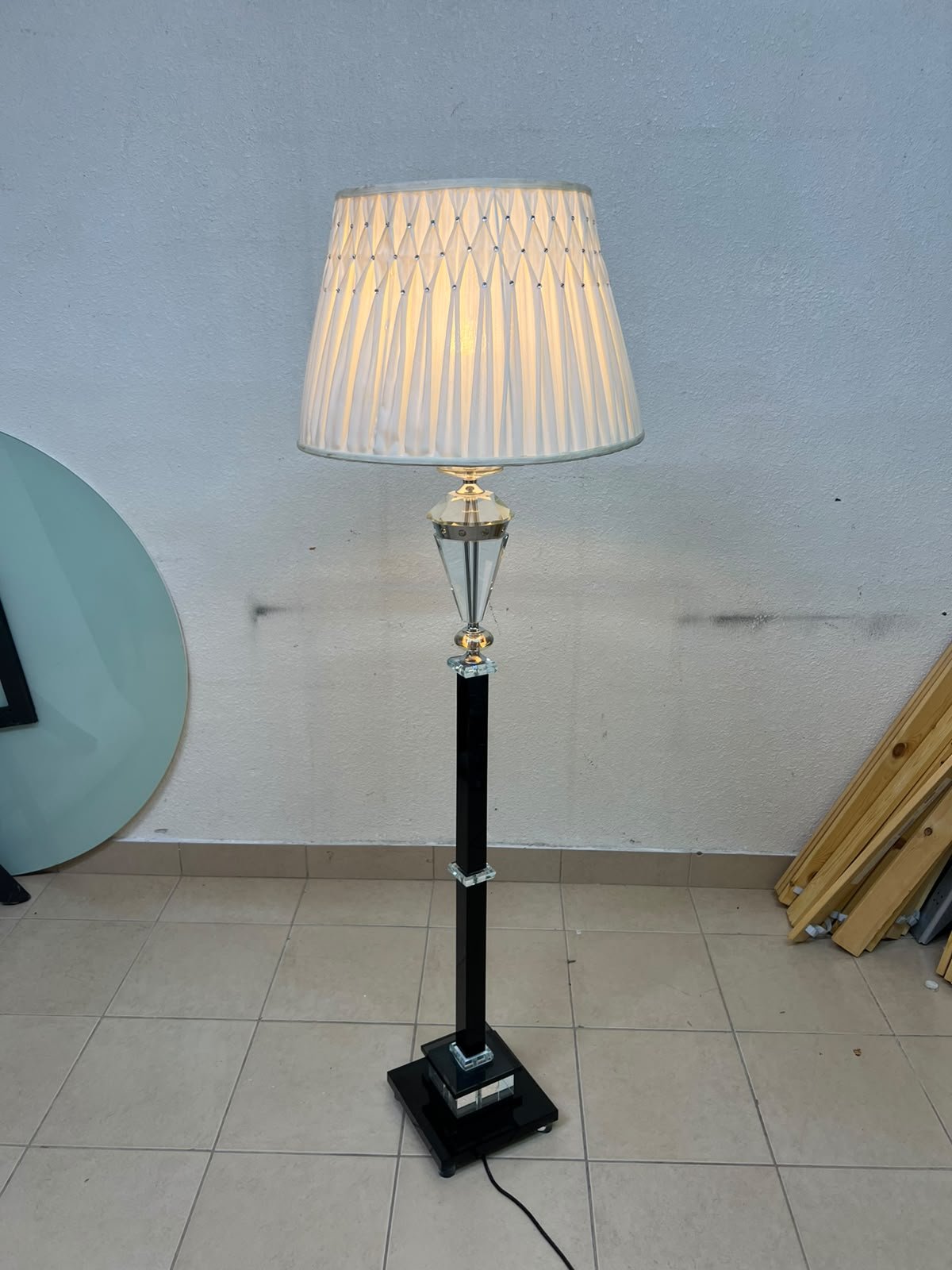 An elegant floor lamp combining crystal and black metal, featuring a conical, pleated fabric shade and a multi-faceted glass element positioned near the top of the lamp's stem.