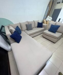 A modern L-shaped corner sofa upholstered in beige fabric, paired with a distinctive standalone lounge chair and accented with contrasting throw pillows in dark blue and patterned gray.