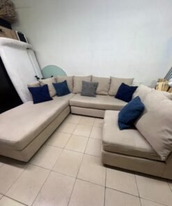 A modern L-shaped corner sofa upholstered in beige fabric, paired with a distinctive standalone lounge chair and accented with contrasting throw pillows in dark blue and patterned gray. - Image 9