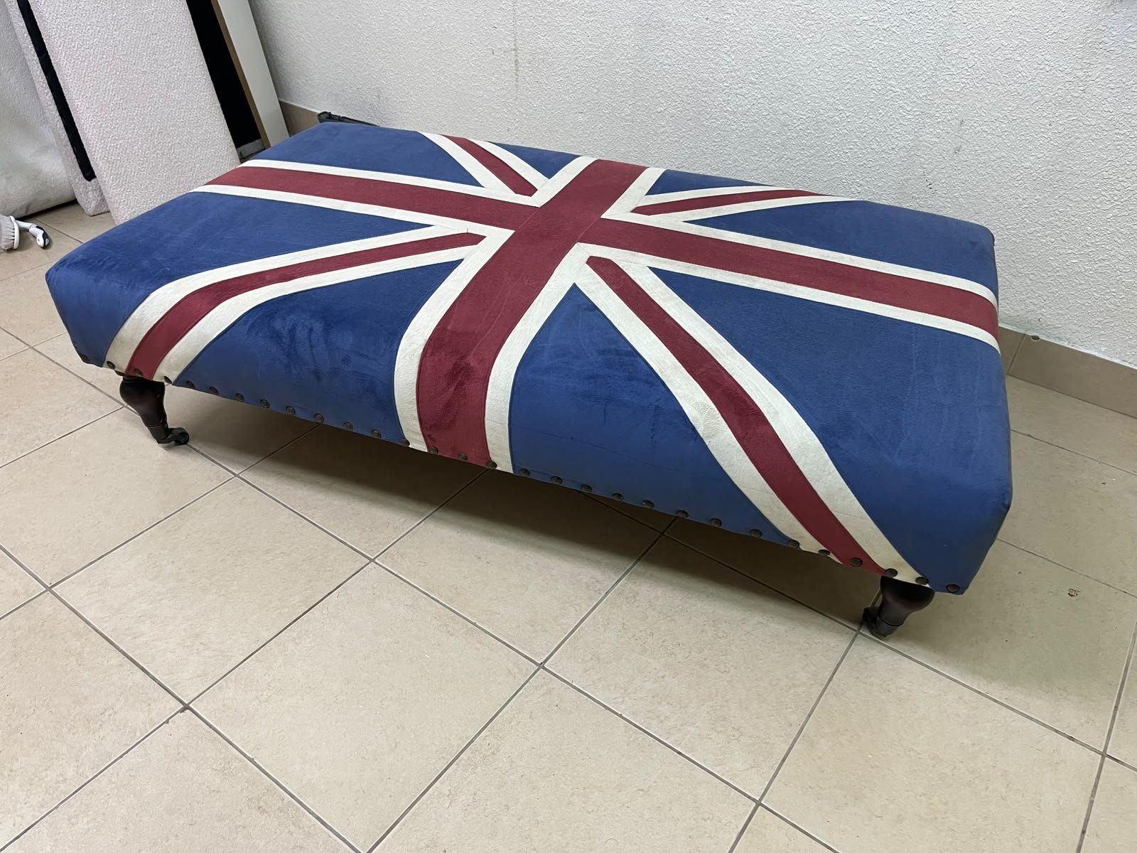 An Ottoman upholstered in luxurious velvet, featuring a vintage British flag design and metal stud detailing along the sides.