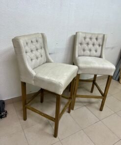 A pair of bar stools featuring a wingback design, beige fabric upholstery with button tufting, and wooden legs. - Image 2