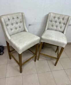 A pair of bar stools featuring a wingback design, beige fabric upholstery with button tufting, and wooden legs.
