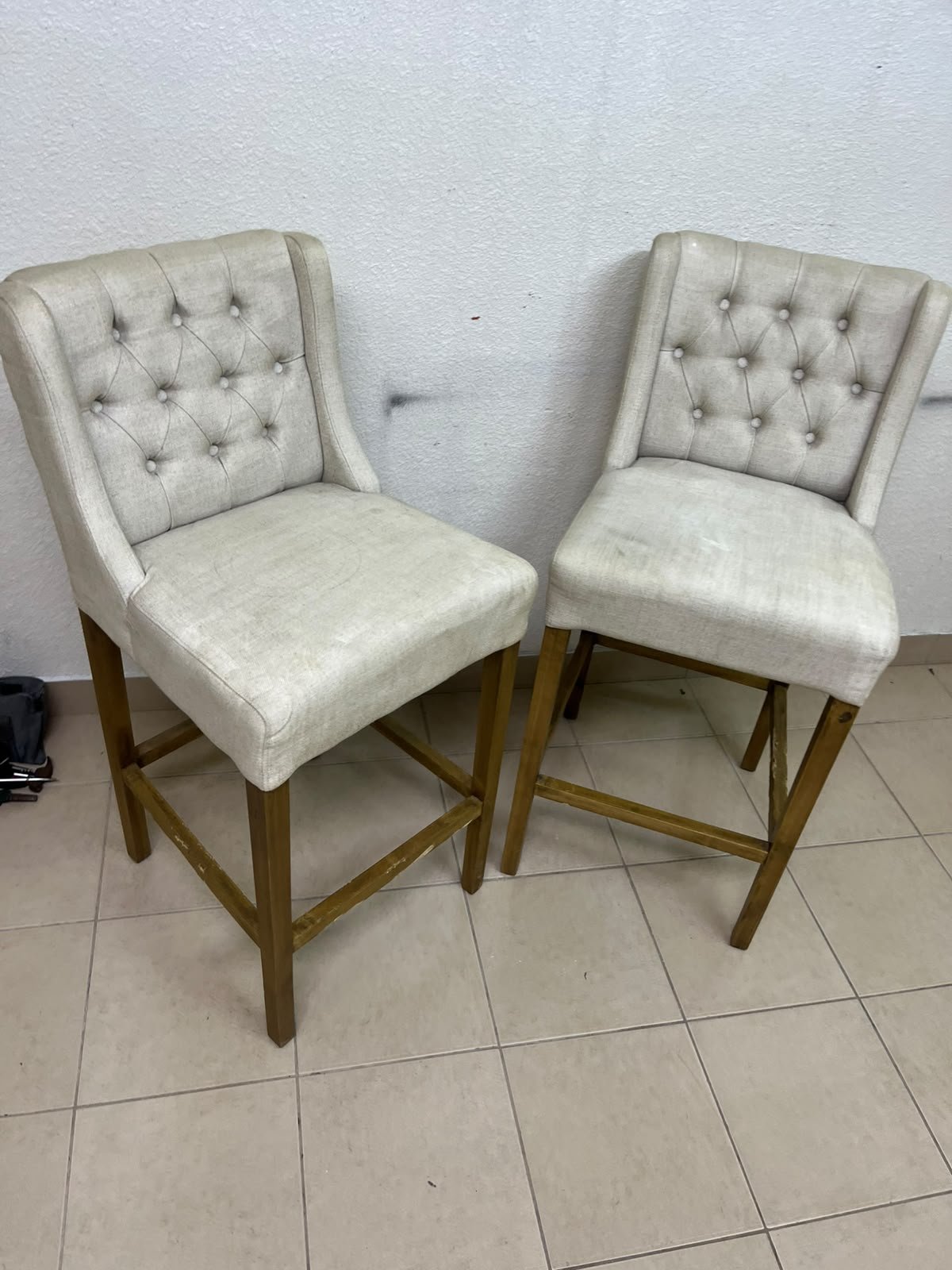 A pair of bar stools featuring a wingback design, beige fabric upholstery with button tufting, and wooden legs.